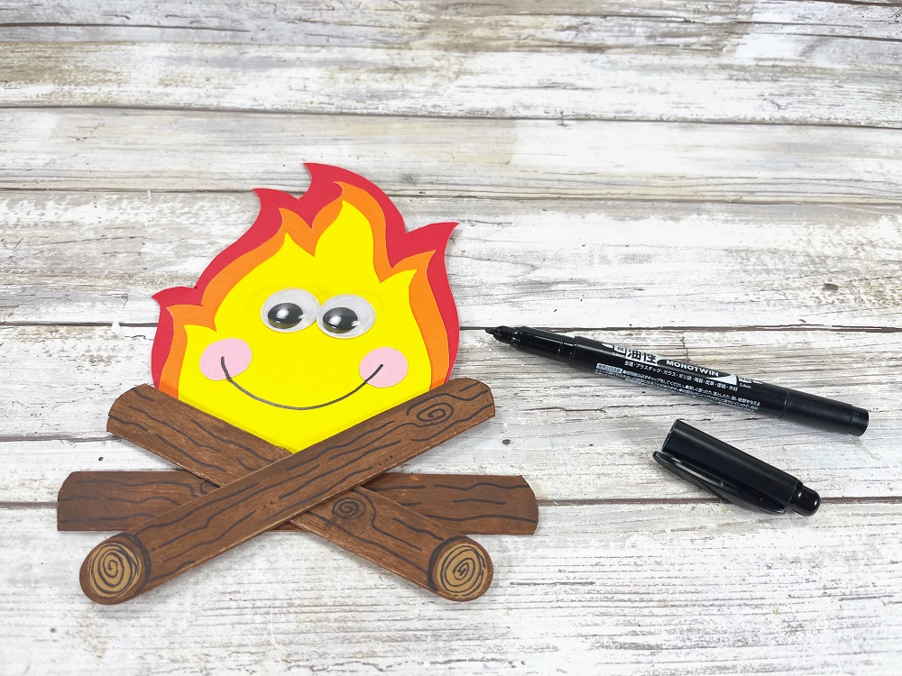 Paper and craft stick campfire craft with google eyes added and kawaii smile.
