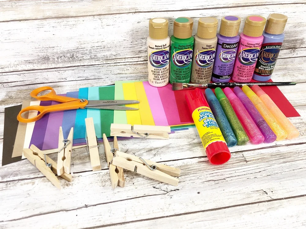 Craft supplies for making clothespin mermaids arranged for photo. Supplies include assortment of colored cardstock paper, wood clothespins, scissors, glue stick, various colors of craft paint in bottles, and several different colors of glitter glue pens.