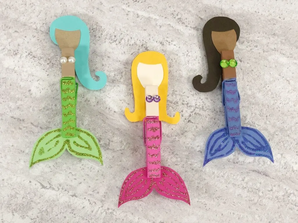 Set of three completed clothespin mermaids arranged next to each other. One has dark skin tone, black hair, and dark blue tail. One has light skin, yellow hair, and pink tail. One has light brown skin tone, aqua hair and lime green tail.