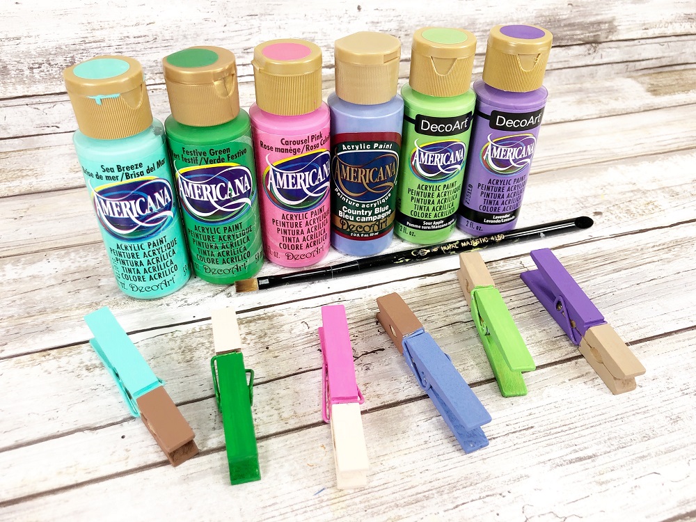 Six different colors of craft paint bottles lined up and a clothespin in front of each one with part of it painted. Colors include aqua, green, pink, blue, lime and purple.
