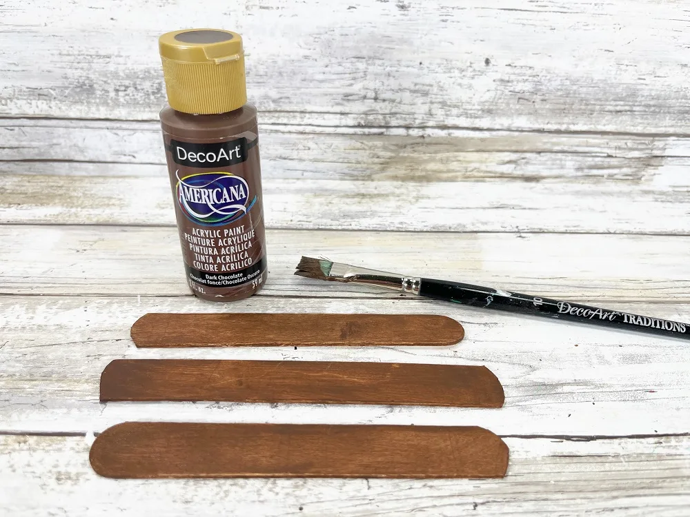 Three popsicle craft sticks painted brown. Sticks are laying near paint brush and bottle of brown paint.