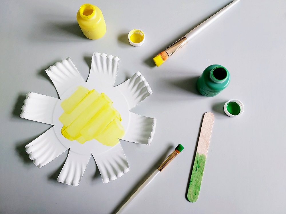 White paper plate cut like a flower being painted yellow. Popsicle craft stick being painted green.