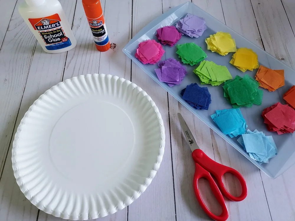 Paper plate, white liquid glue bottle, glue stick, scissors, and tray with nicely arranged tissue paper squares sorted by color are crafting supplies all arranged on table.