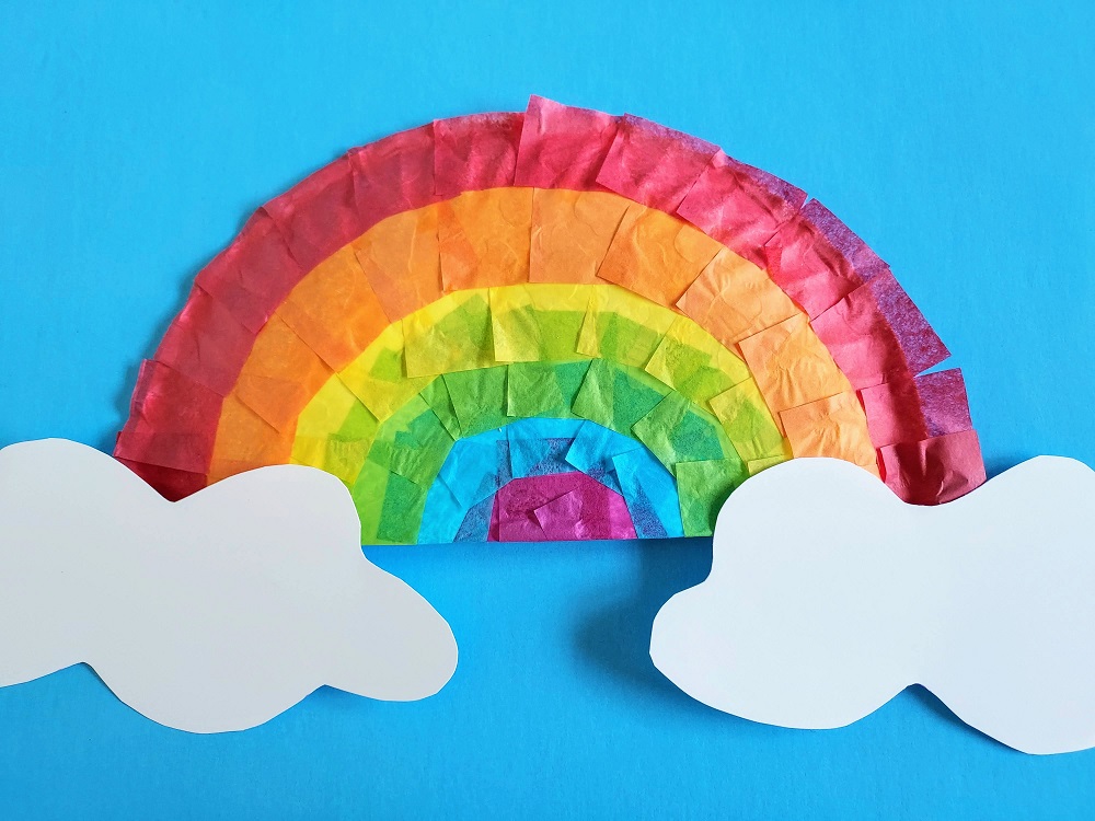 Close view of completed rainbow craft made with tissue paper and a paper plate. White clouds are attached to the plate. Craft is laying on blue paper to look like the sky.