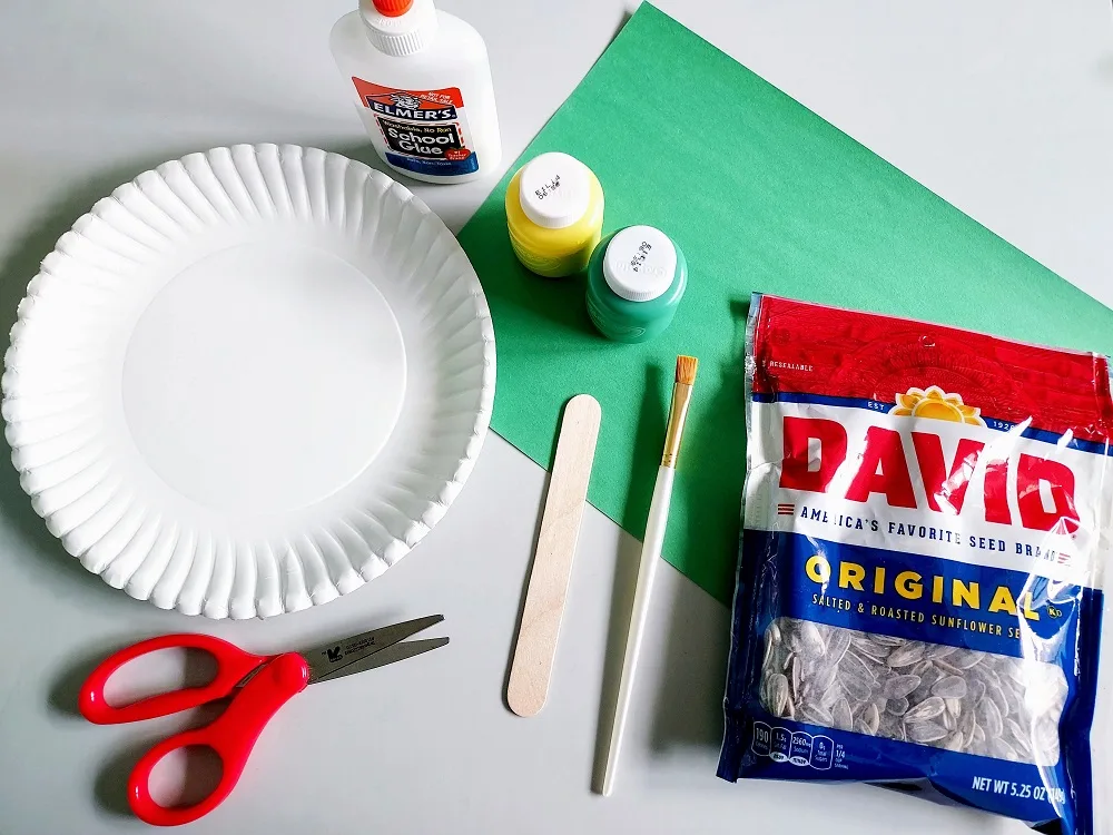 Craft supplies arranged on table to make paper plate sunflower. Materials include paper plate, scissors, glue, yellow paint, green paint, green construction paper, popsicle craft stick, paint brush, and bag of sunflower seeds.