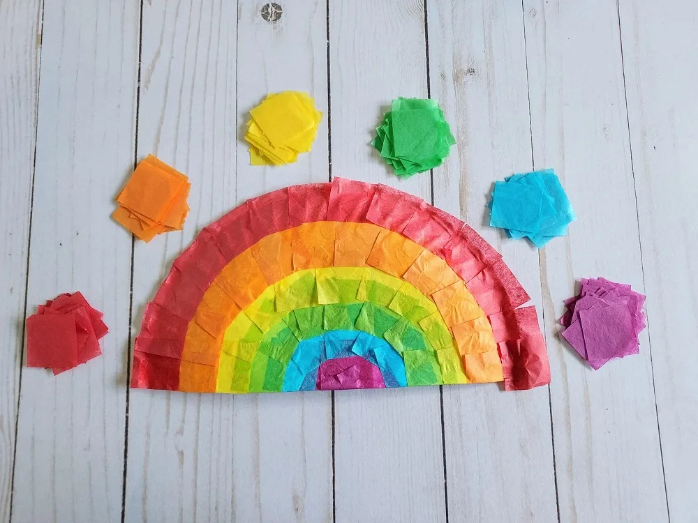 Paper plate covered with tissue paper squares in ROYGBIV rainbow order. Stacks of tissue paper squares arranged in an arch above completed craft.