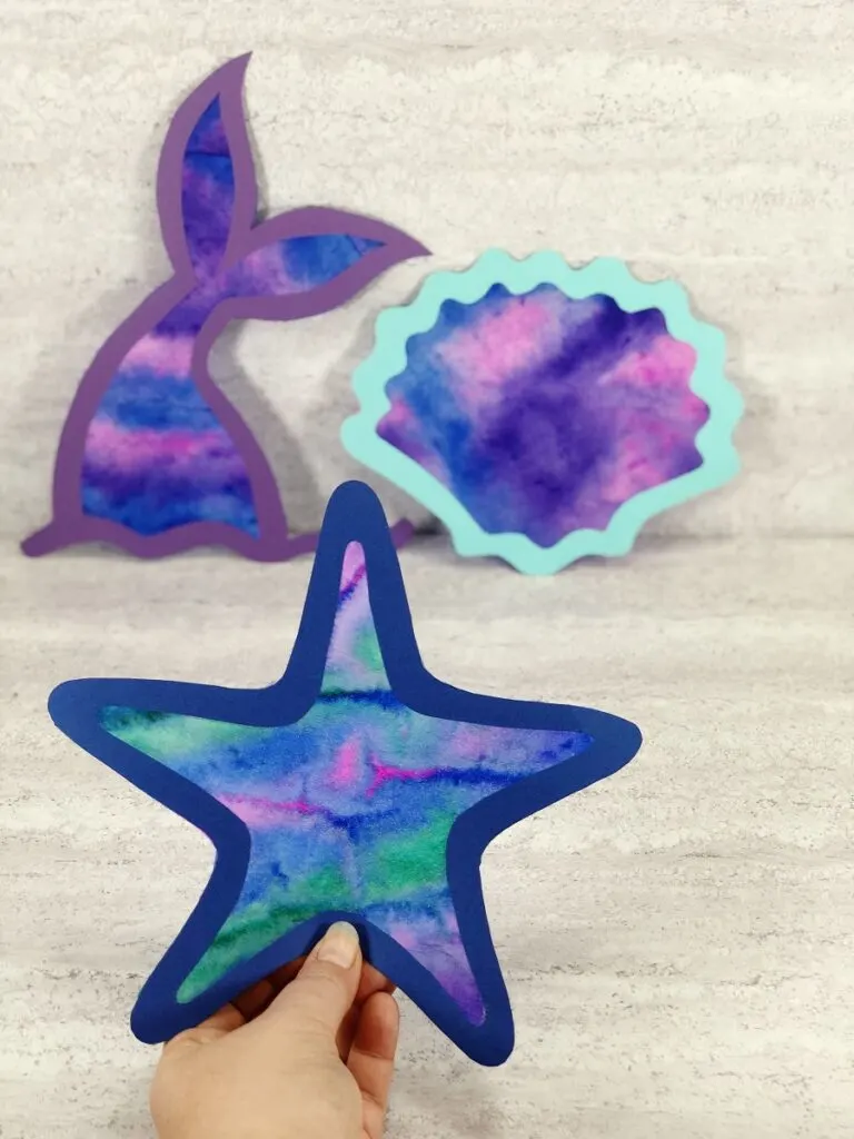 Set of three completed coffee filter suncatcher crafts. Hand holding up starfish and mermaid tail and seashell in the background.