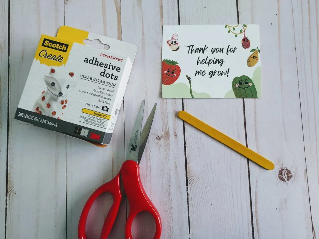 Box of glue dots, a pair of scissors, yellow colored popsicle stick, and cut out plant themed gift tag laying out on table.