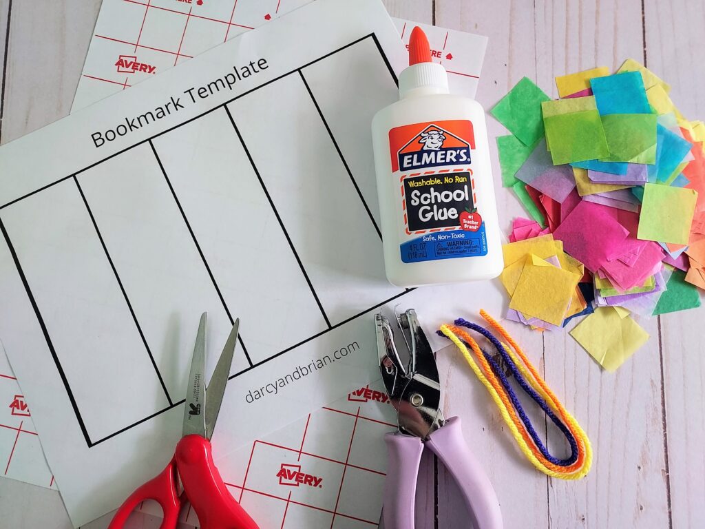 Bookmark craft supplies laying on table includes printed out blank bookmark template, scissors, hole punch, white school glue, pieces of yarn, and square pieces of tissue paper in a variety of bright colors.