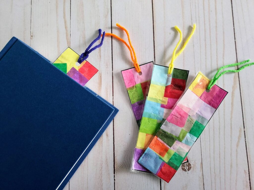 Overhead view of one tissue paper bookmark sticking out of a closed book laying next to three more finished bookmarks.