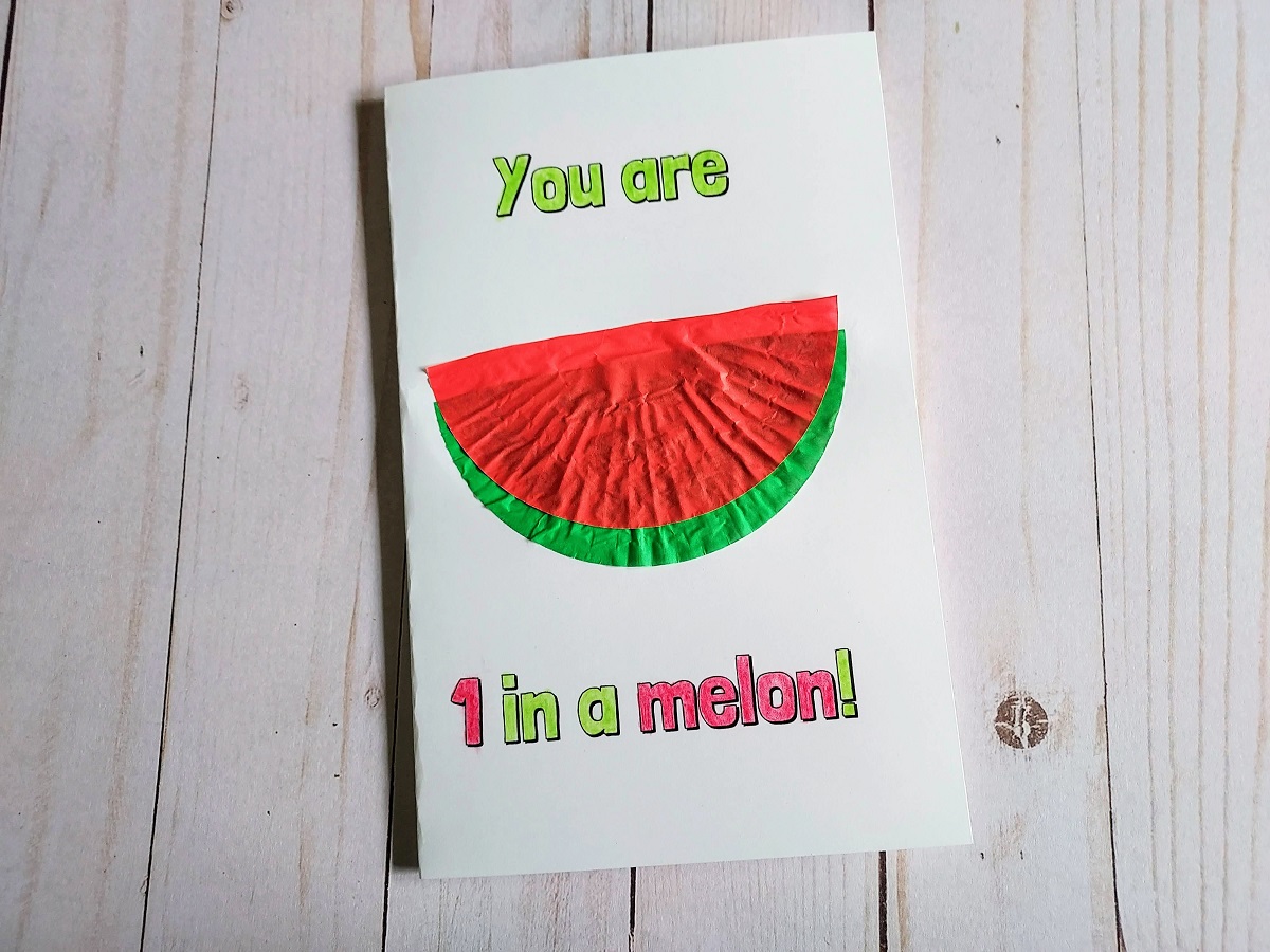 Half of a flattened red cupcake liner has been glued offset on top of a green liner to look like a slice of watermelon on the front of a handmade card.