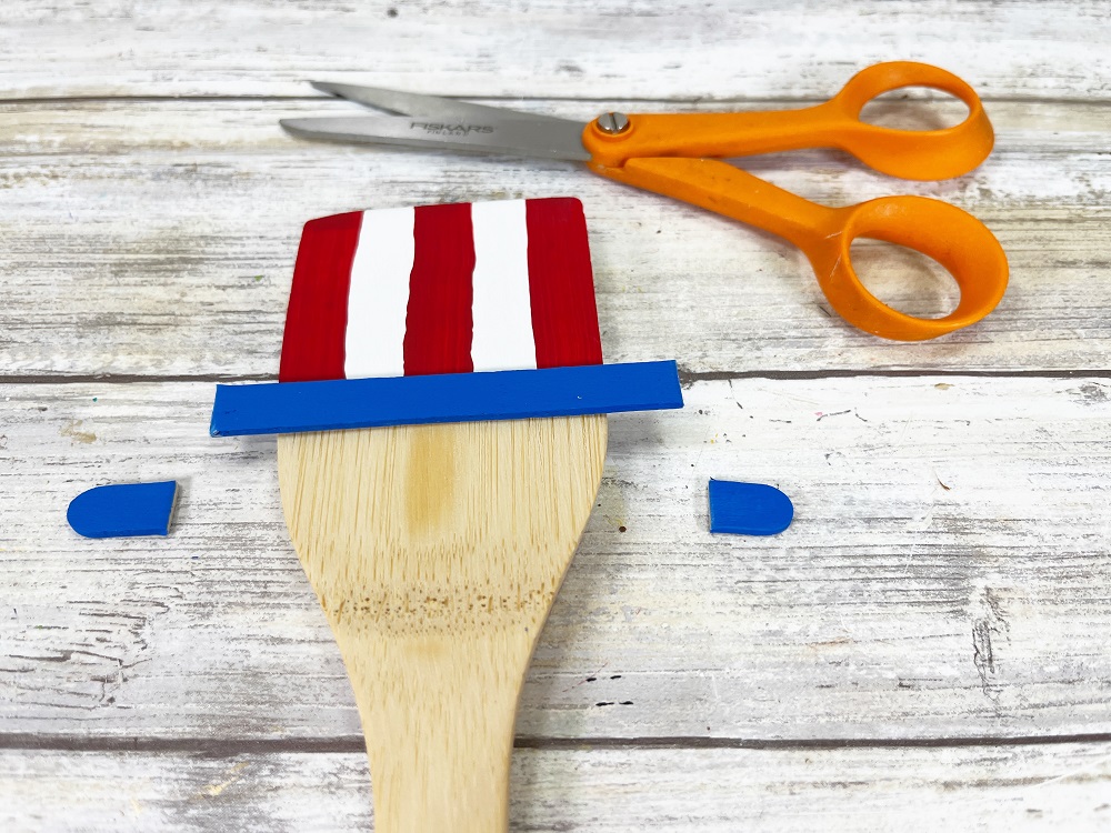 Close view of top part of bamboo spatula. Top section is painted in red and white vertical stripes. Blue painted craft stick has been glued on horizontally to create hat brim. The ends of the craft stick have been trimmed.