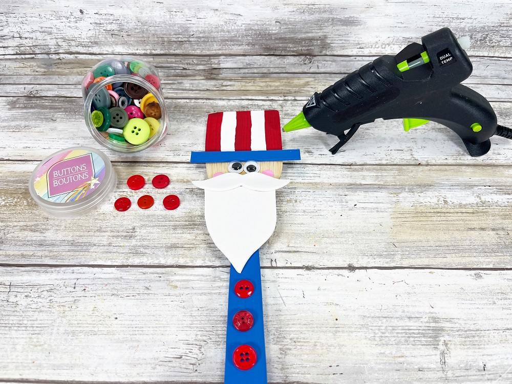 Three red buttons added onto handle below the beard. Uncle Sam craft almost completed. Glue gun and container of assorted colors of buttons are next to project.