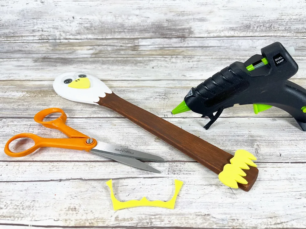 Wooden spoon painted to look like a bald eagle with yellow craft foam claws glued to the handle. Glue gun, scissors, and scrap of craft foam lay around the project.