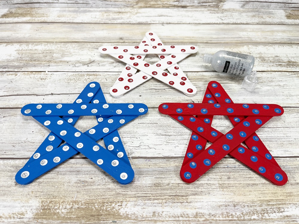 Small bottle of silver glitter glue laying near popsicle stick stars. There are three arranged in a triangle: painted blue with white polka dots, white with red polka dots, and red with blue dots. Silver glitter glue added to center of dots.