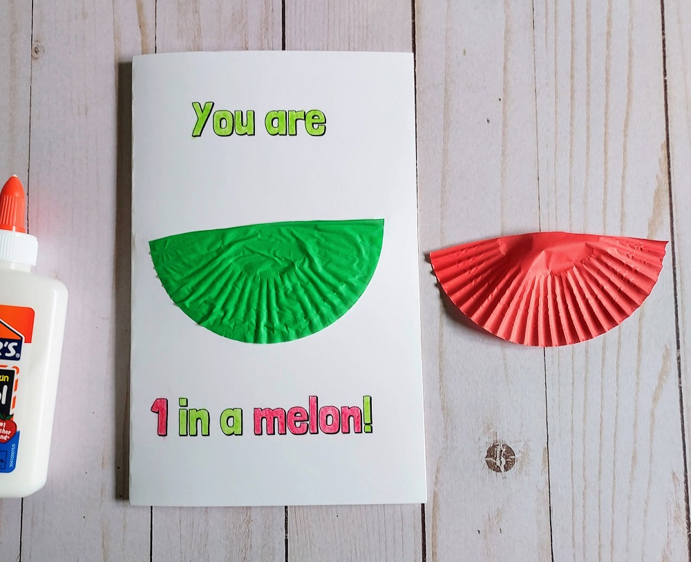 Half of a flattened green cupcake liner is glued to the center of a printable card with a melon pun. Half of a red baking cup lays next to it as does a bottle of school glue.