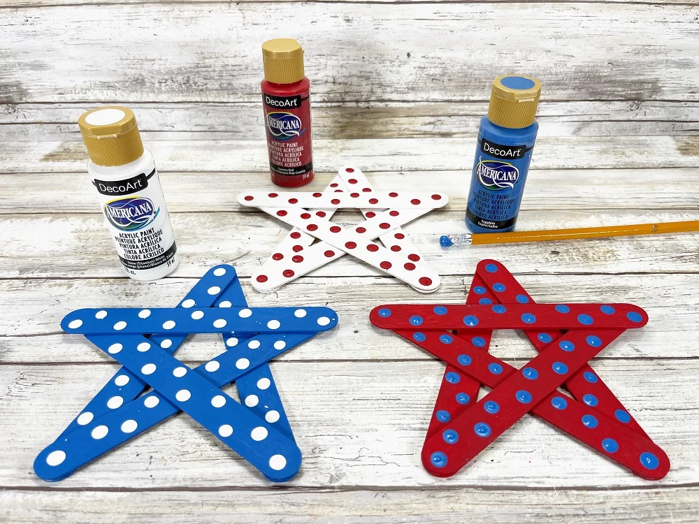 From left to right: blue painted popsicle stick star with white dots is in front of bottle of white paint. White painted star has red dots and is next to red paint bottle. Star painted red has blue polka dots and is laying in front of blue paint and pencil with paint on the eraser end.