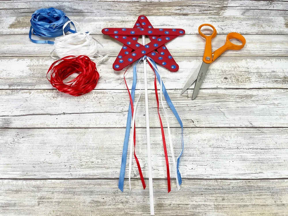 Red painted star with blue polka dots attached to white dowel. Thin red, white, and blue ribbons tied on under bottom of star. Pair of scissors and three piles of ribbon lay nearby.
