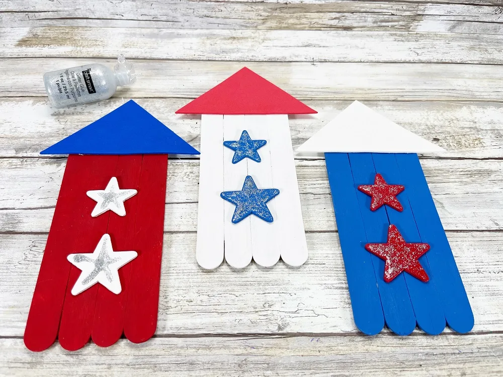 Three in progress craft stick firework rockets. Left one has blue triangle top glued onto red painted craft sticks with white stars. Middle one has red top, white base, and blue stars. Left rocket has blue base with red stars and white topper.