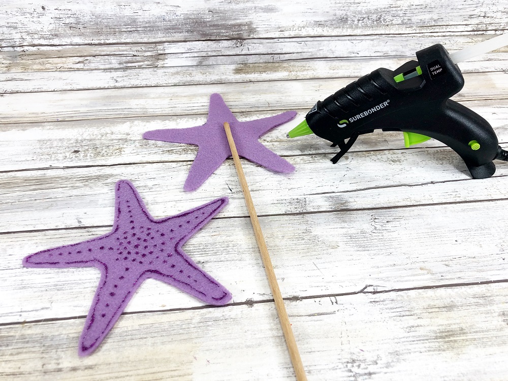 One purple craft felt starfish with wooden dowel glued to the middle of it. Glue gun sitting to the right of them. To the left is the purple felt starfish decorated with glitter glue waiting to be attached to the wand.