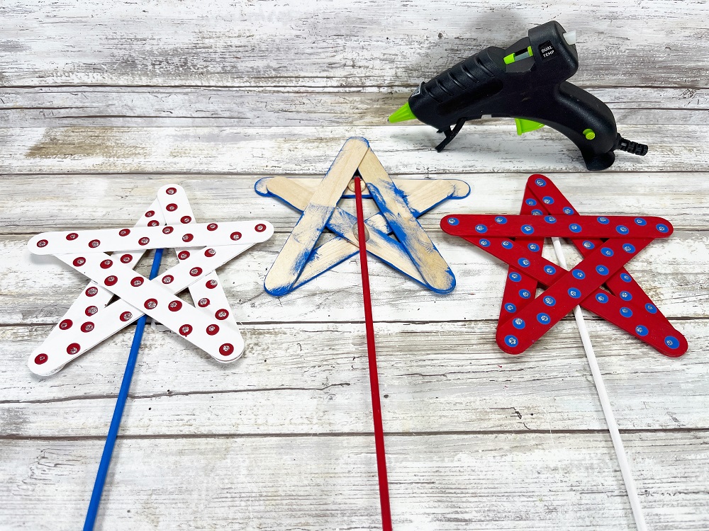 Three star wands laying side by side. Center star is turned over (paint side down) to show where the red painted dowel was glued in place. Wooden dowels already attached to the other two.