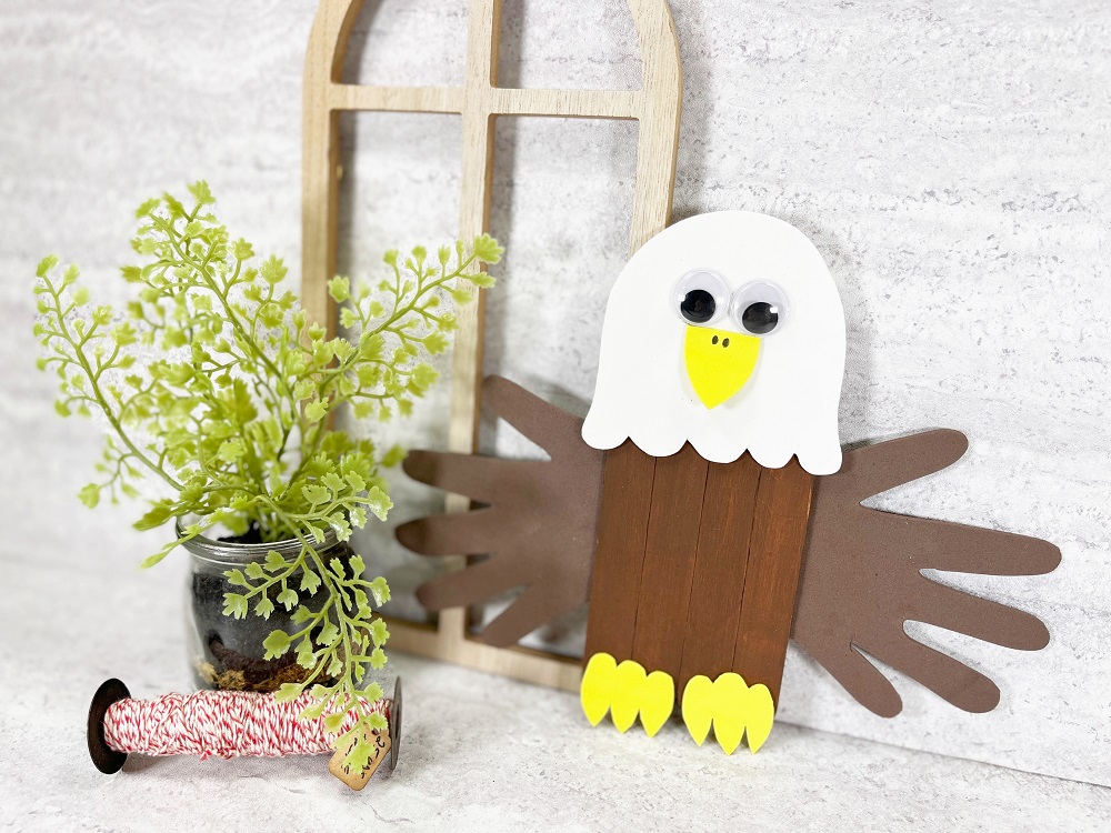 Completed bald eagle craft stick project leaning against wooden frame and light gray backdrop. Small green plant and spool of red and white twine are on the left.