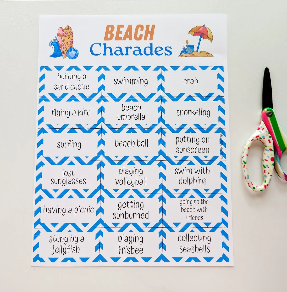 One page of beach charades printed out and laying on white desk top next to a colorful pair of kids scissors.