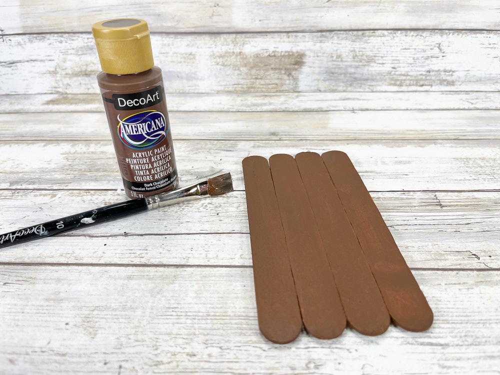 Bottle of brown paint and paint brush lay to the left of brown painted craft sticks.