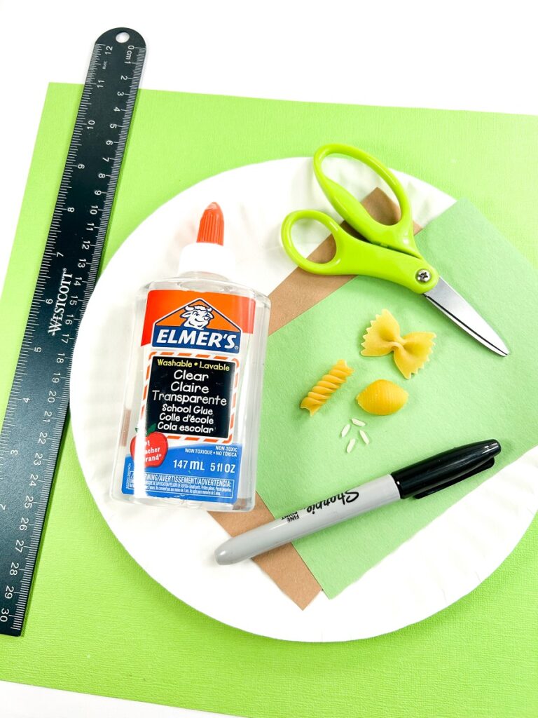 Bottle of glue, different types of dry pasta, some grains of white rice, a black marker, scissors, and green and brown construction paper are on top of a paper plate. The paper plate is on top of a larger bright green piece of paper with a ruler next to it.