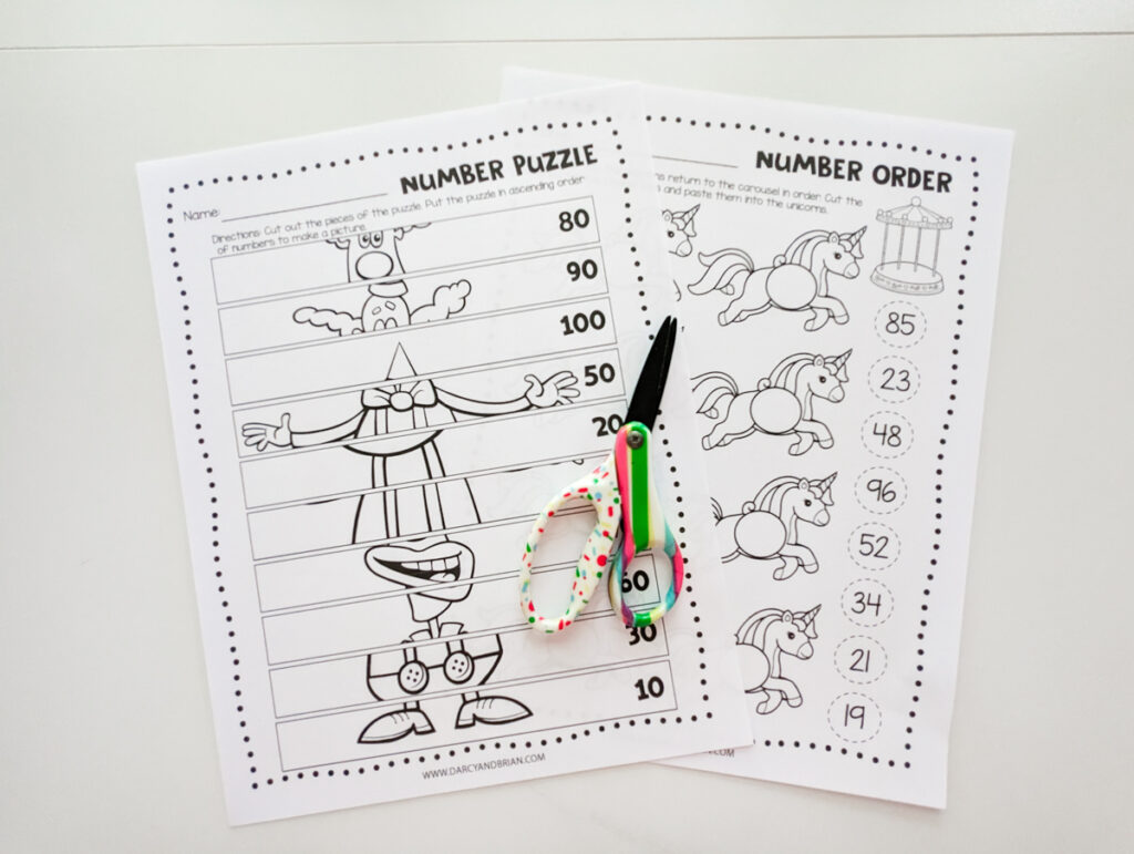 Two worksheets with cut and paste activities printed out and laying overlapping on white desk. Top page shows a clown number order picture puzzle. Colorful kid scissors lay on top of the papers.