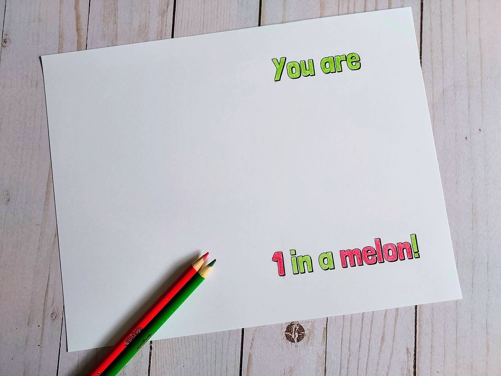 Printable melon themed card laying open on table. The words "You are 1 in a melon!" have been colored in. Red and green colored pencils lay on the card.
