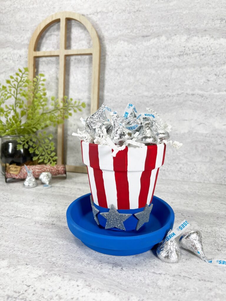 Clay pot painted to look like a patriotic hat with red and white stripes, silver stars, and a blue painted saucer as the hat brim. Flower pot is filled with wrapped candy.