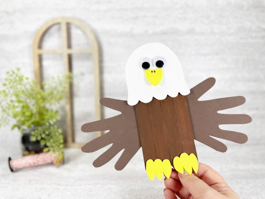 Caucasian woman's hand holding up a completed bald eagle craft made with jumbo popsicle sticks and handprint wings. Small wooden arch frame and small green plant are in the background.