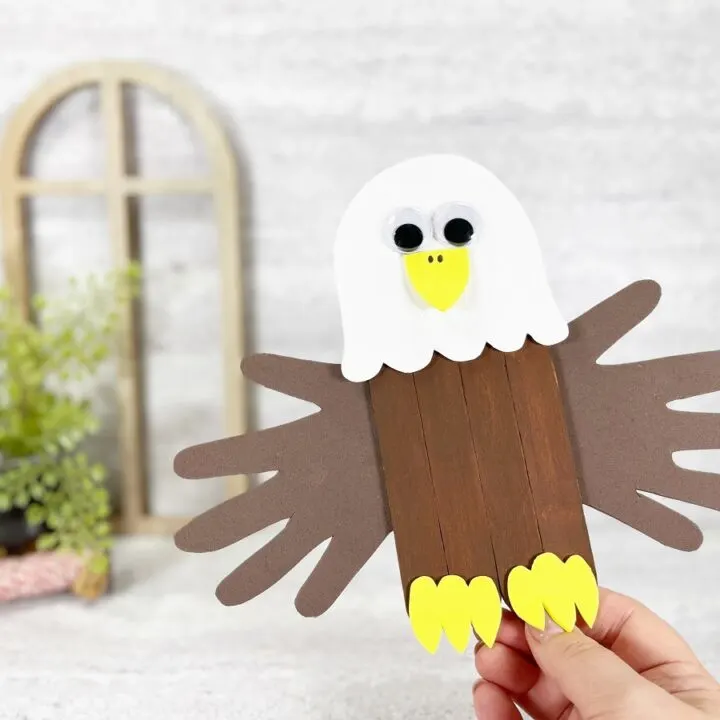 Caucasian woman's hand holding up a completed bald eagle craft made with jumbo popsicle sticks and handprint wings. Small wooden arch frame and small green plant are in the background.