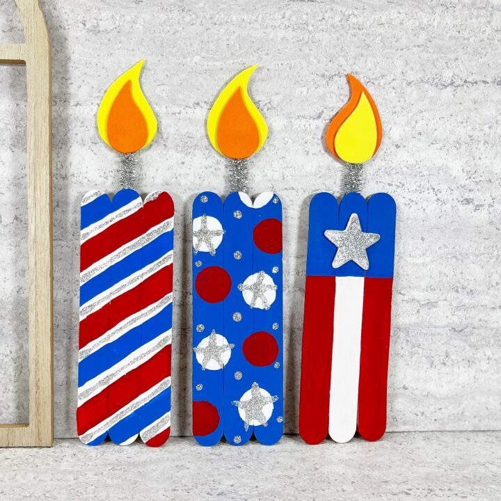 Three completed firecracker crafts made with jumbo craft sticks. Each one is painted in a different design using red, white, and blue paints. One is striped, another has polka dots, and another is flag inspired.