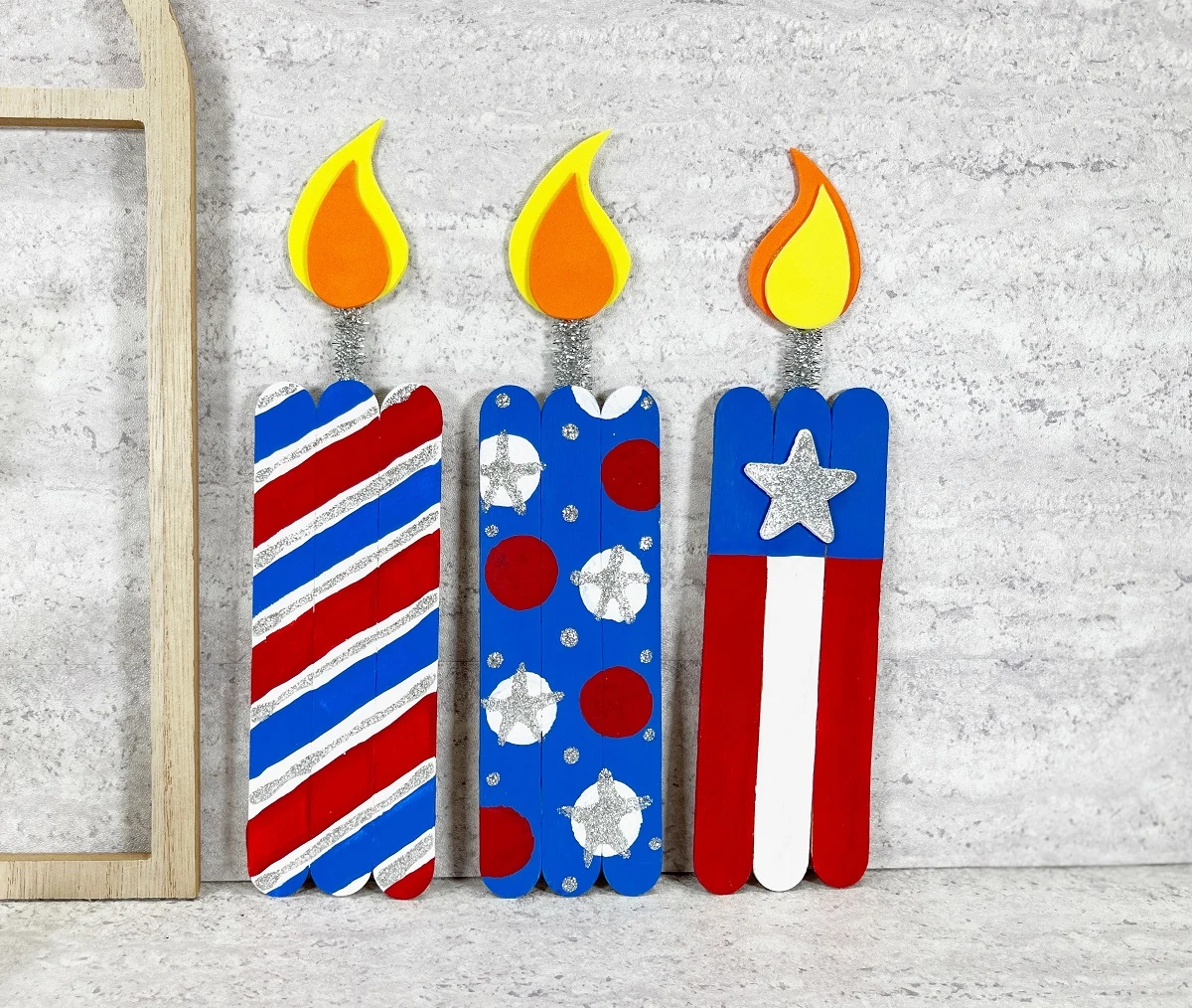 Three completed firecracker crafts made with jumbo craft sticks. Each one is painted in a different design using red, white, and blue paints. One is striped, another has polka dots, and another is flag inspired.