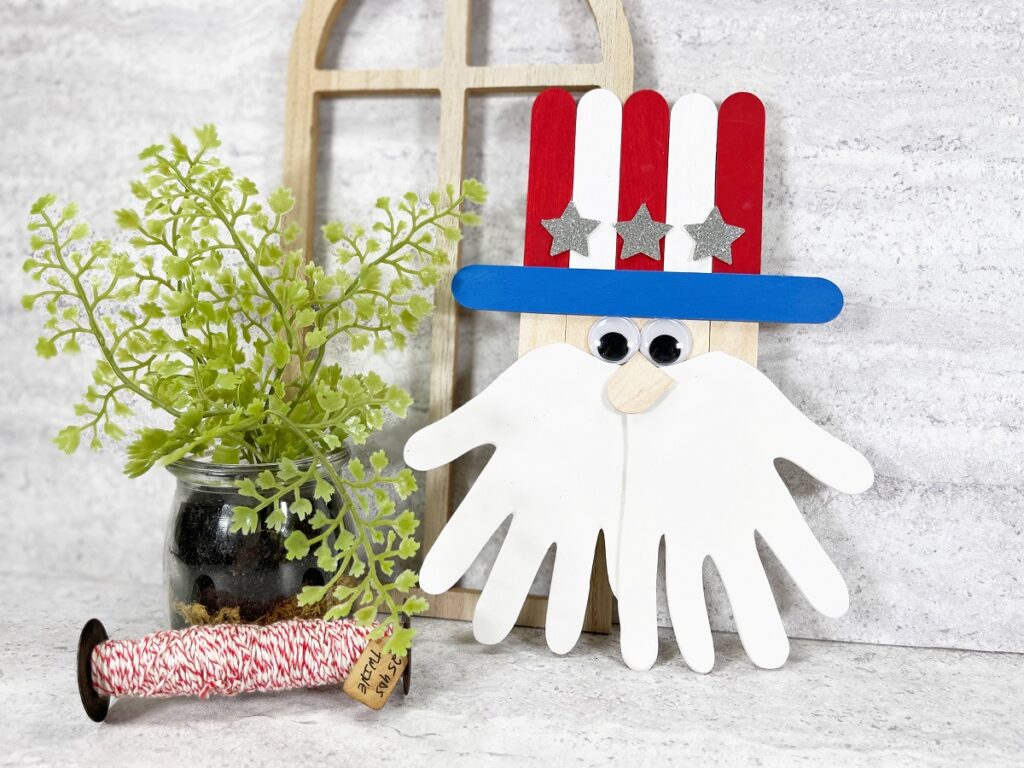 Uncle Sam popsicle stick craft completed finished and leaning up against the wall by a wooden frame and small plant. Craft made with red, white, and blue painted popsicle sticks and two white handprint pieces of craft foam for a beard.