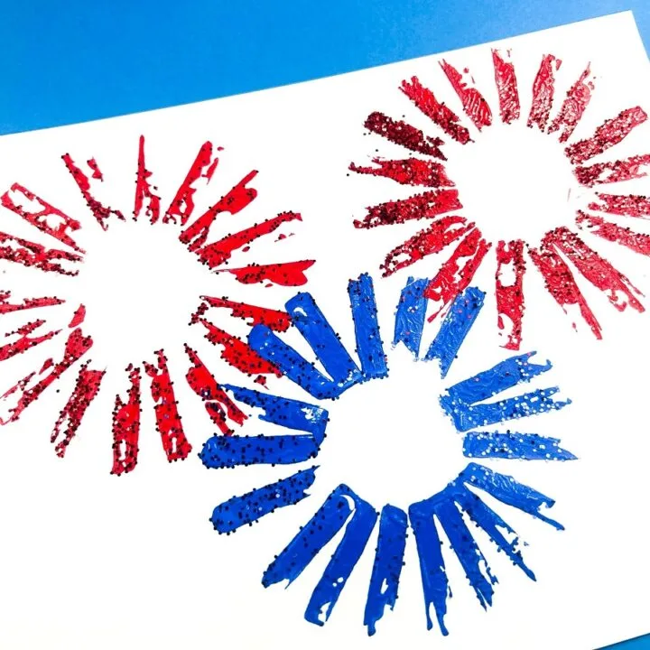 Overhead view of white cardstock paper in landscape orientation on a blue background. Red and blue paint stamped fireworks on the white paper with glitter.
