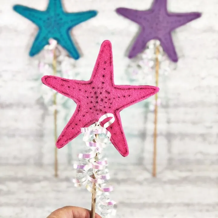 Caucasian woman's hand holds up pink mermaid wand in the foreground. Blurred in the background are two more wands. A blue one and a purple one.