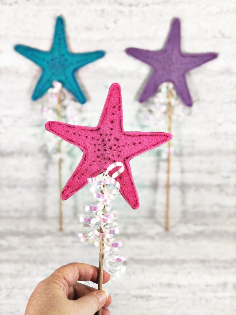 Caucasian woman's hand holds up pink mermaid wand in the foreground. Blurred in the background are two more wands. A blue one and a purple one.