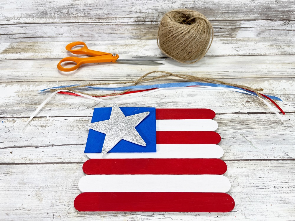 Assembled popsicle stick flag with ribbon, twine, and scissors laying along the top, waiting to be attached to the back.