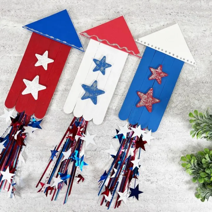 Three red, white, and blue painted popsicle stick fireworks rockets side by side and angled up towards the right. Small green plant on the right side.