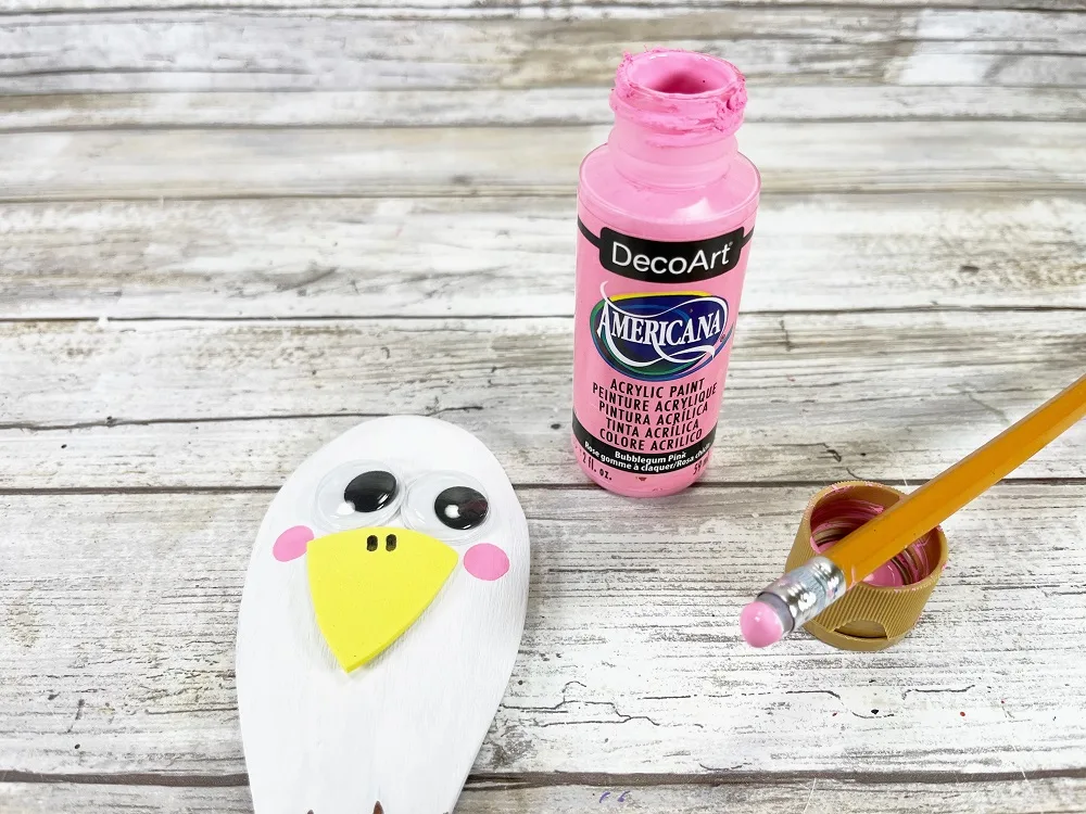 Close view of the spoon head which is painted white. Two googly eyes and a yellow beak have been glued to the back of the spoon. Pencil with eraser covered in pink paint laying next to eagle which has two pink cheeks from dotting the paint covered eraser.