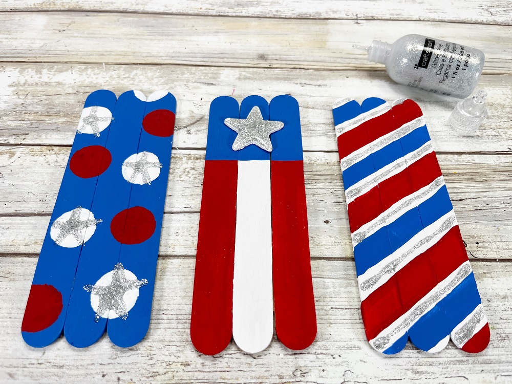 Three different craft stick firecracker bases. Left one is blue with white and red dots. Middle one is decorated like the American flag. Right one has diagonal red, white, and blue stripes. The white is accented with silver glitter glue.