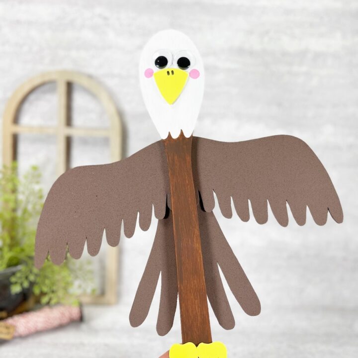 White woman's hand holding up the wooden spoon Bald Eagle craft project. Brown craft foam wings and tail feathers have been glued to the back.