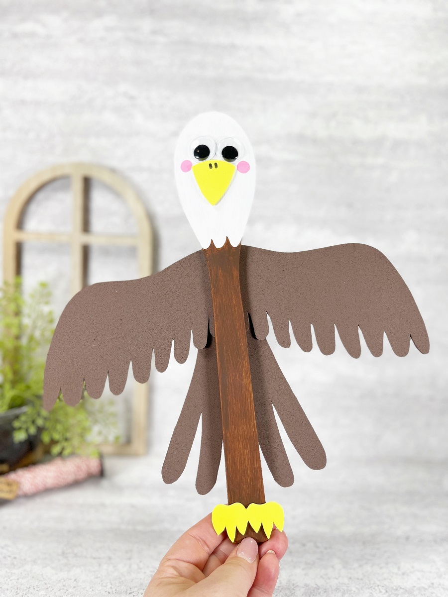 White woman's hand holding up the wooden spoon Bald Eagle craft project. Brown craft foam wings and tail feathers have been glued to the back.