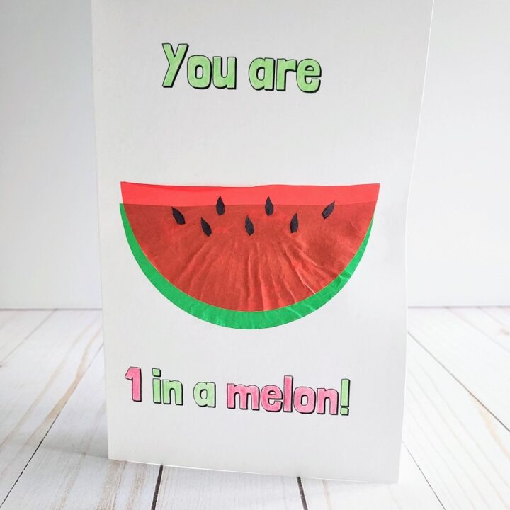 Completed card craft standing up. Text on cared is colored in green and red. It says You are 1 in a melon! A slice of watermelon made using paper baking cups is in the center on the front of the card.