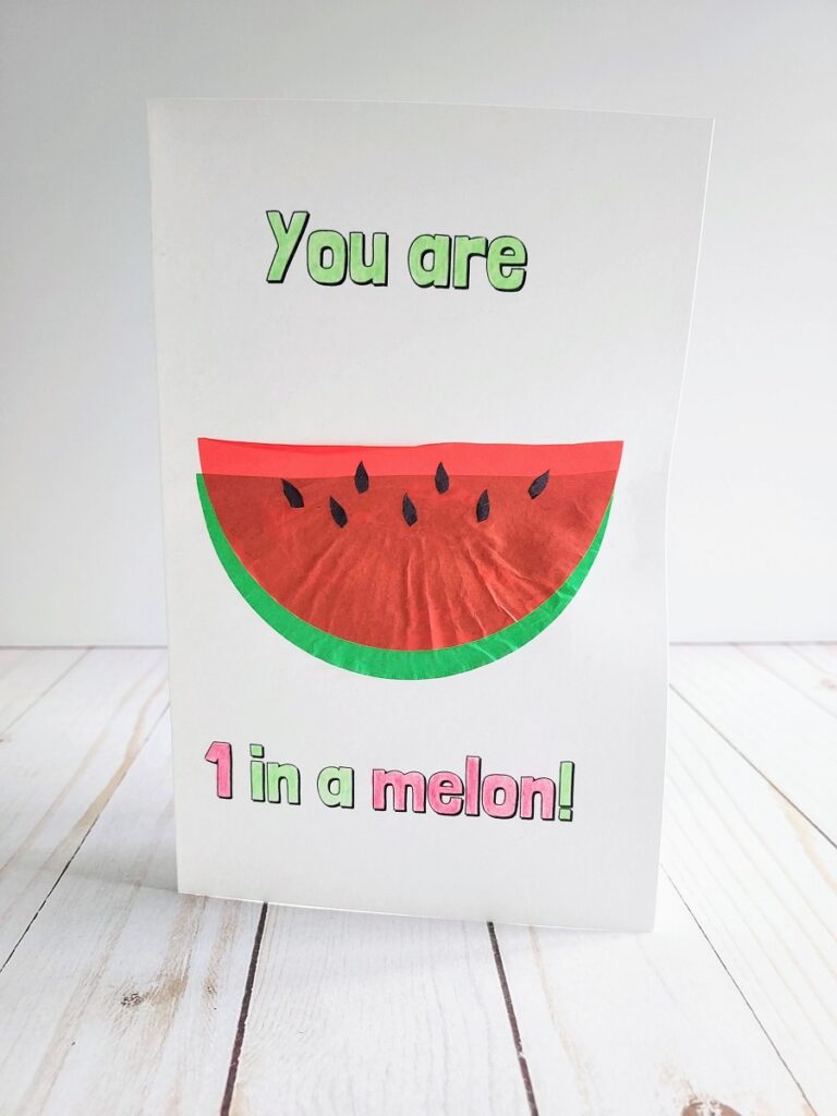 Completed card craft standing up. Text on cared is colored in green and red. It says You are 1 in a melon! A slice of watermelon made using paper baking cups is in the center on the front of the card.