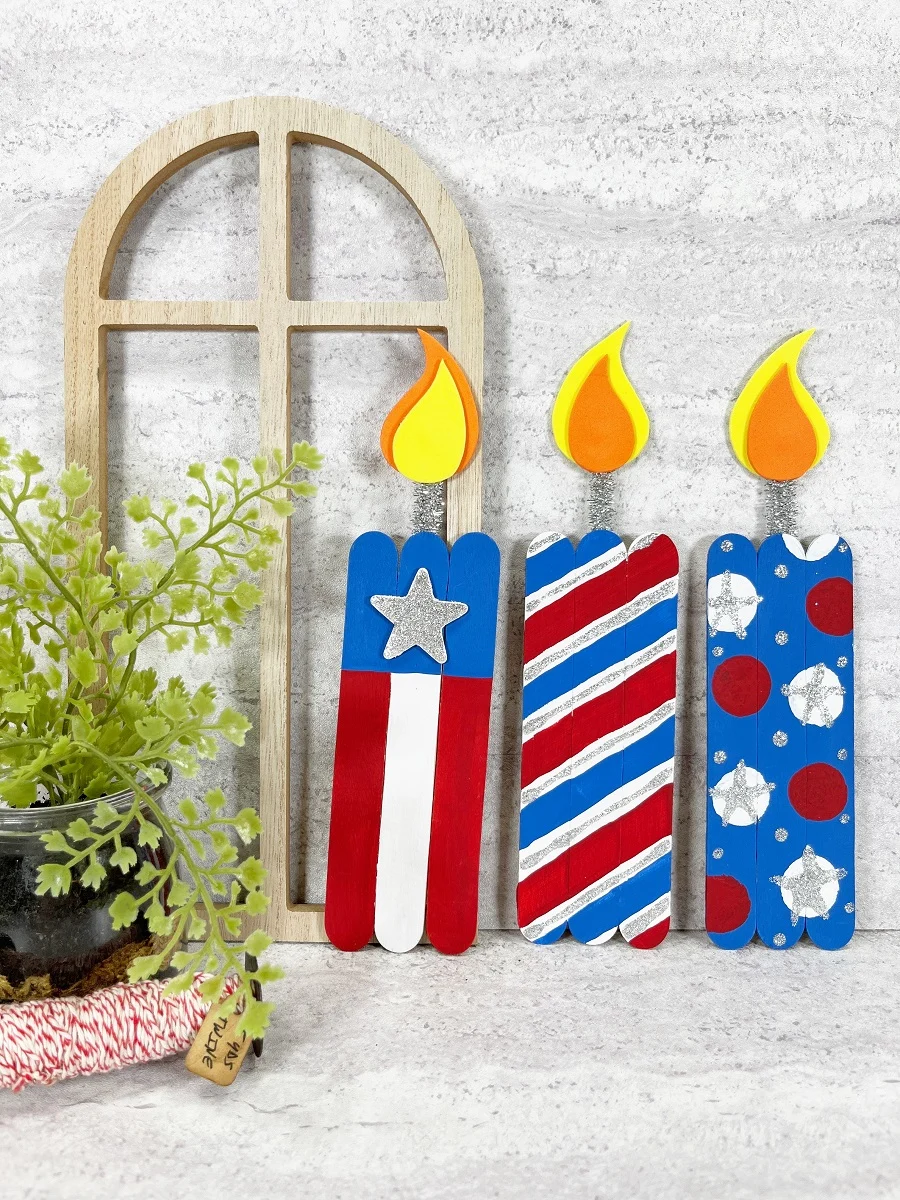 Three completely finished and assembled popsicle stick firecrackers lined up next to each other leaning against the wall. They are made with craft sticks, chenille stems, craft foam, and paint.