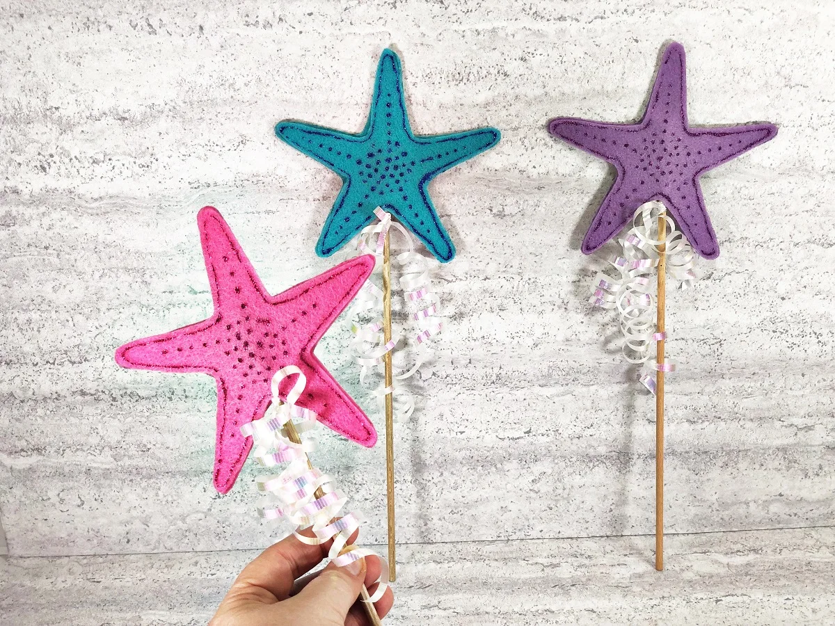 Left hand of Caucasian woman holding up finished mermaid wand craft with a pink felt starfish topper. A blue wand and purple wand are propped up in the background. 